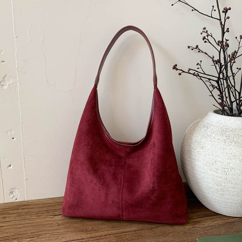 A burgundy suede hobo shoulder bag with a single strap, standing on a wooden surface next to a white textured ceramic vase with dried branches.