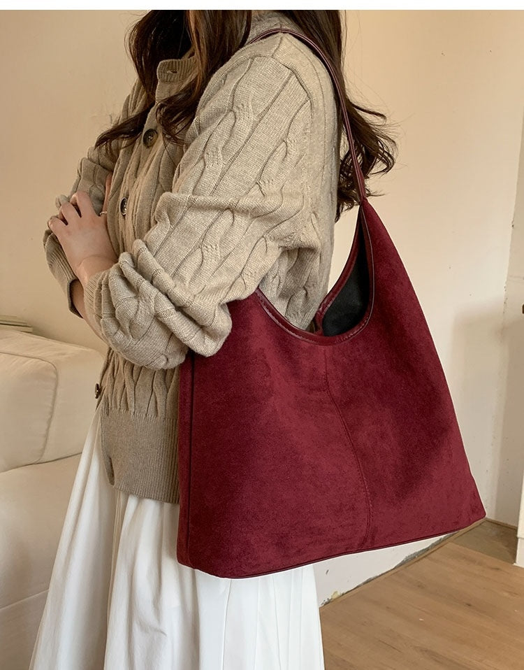 A person wearing a beige cable-knit cardigan and white skirt carries a large slouchy shoulder bag in burgundy suede-like material, shown in an indoor setting.